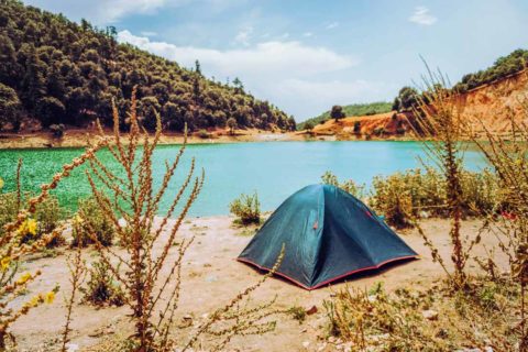 Tent pitched by a lake with green water, hills, and trees in the background