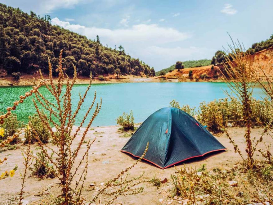 Tent pitched by a lake with green water, hills, and trees in the background